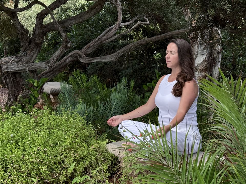 Daiana meditating outdoors in a lush garden, sitting cross-legged with eyes closed and hands resting on her knees.
