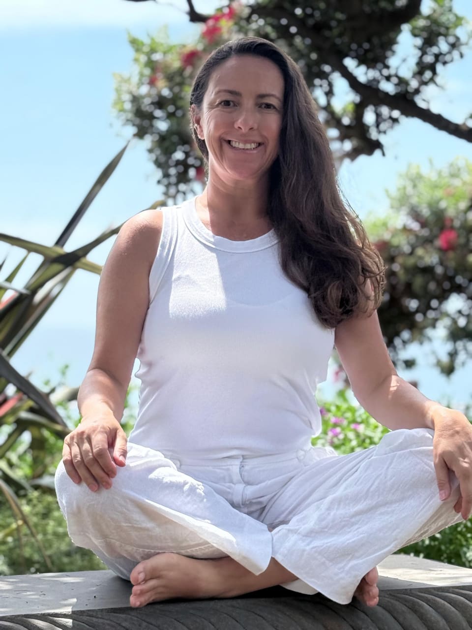 Daiana, with long dark hair sitting cross-legged outdoors on a log, smiling, with a background of trees and flowering plants on a sunny day.