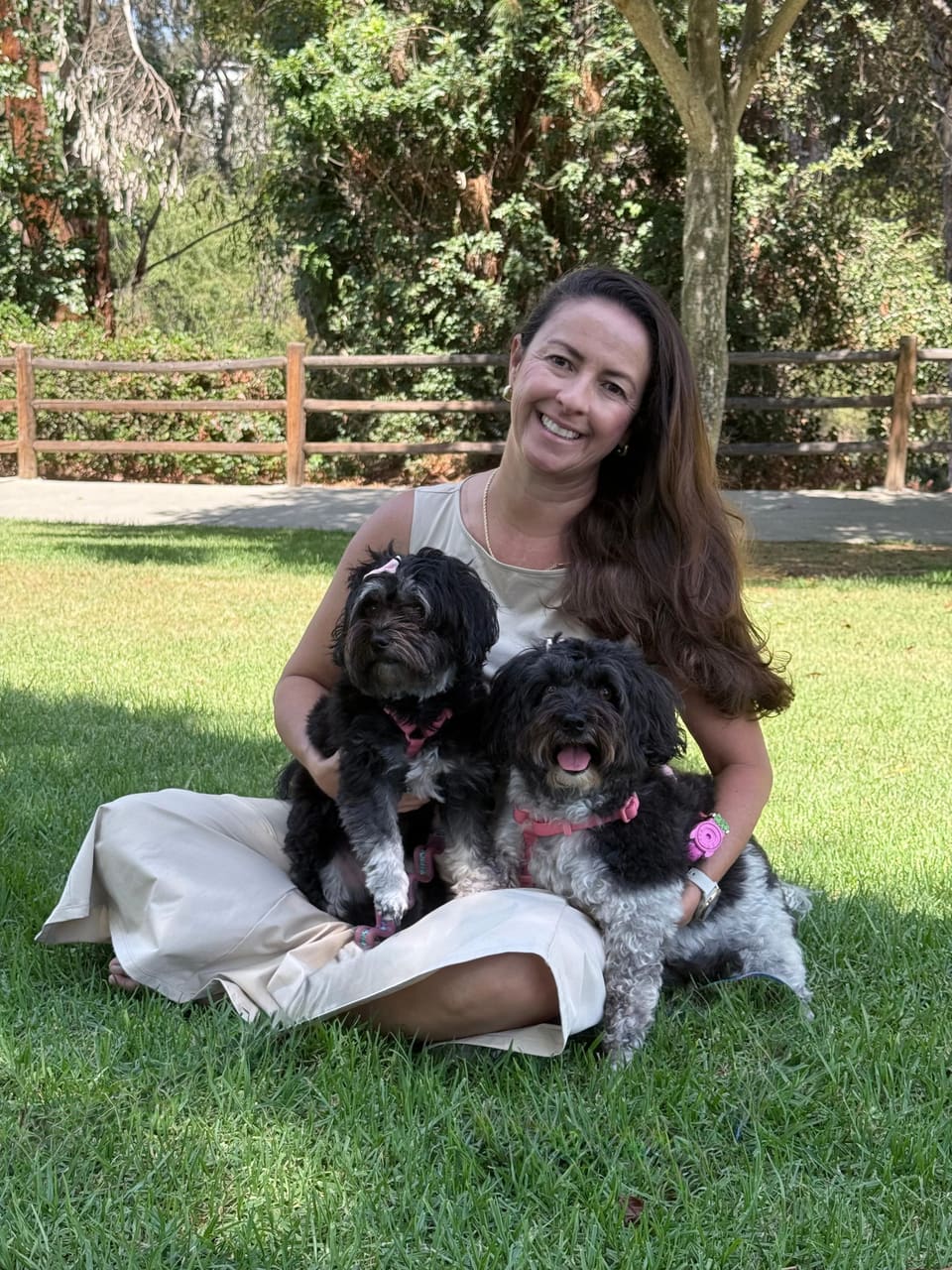 Daiana sitting cross-legged on grass, smiling, holding two small black dogs with white markings, in a park with trees and a wooden fence in the background.